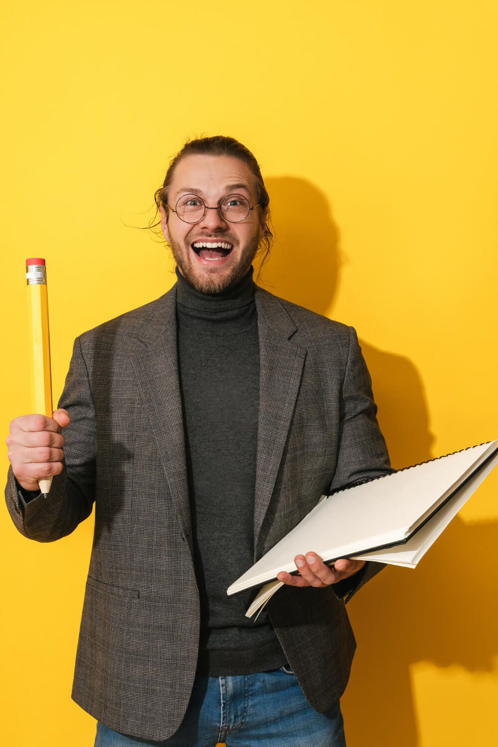 Studente sorridente con quaderno e matita gigante su sfondo giallo, copertina per articolo su come scegliere il relatore giusto per la tesi