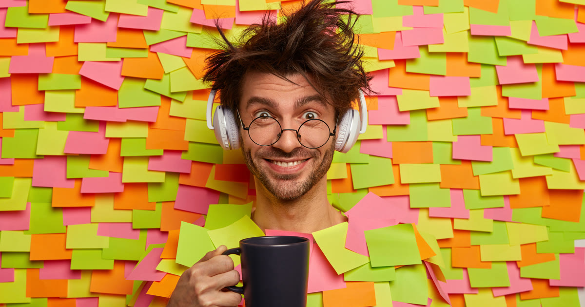 Studente sommerso da post-it colorati che sorride con una tazza in mano: quando non riesci a scrivere la tesi si riparte da compiti piccoli